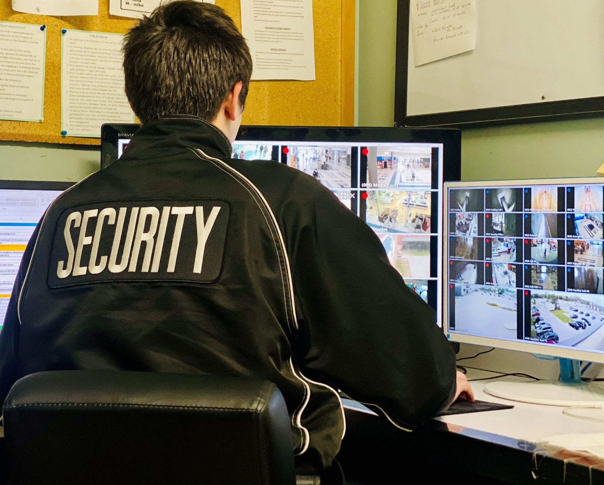 Security guard sitting in an office watching video surveillance footage on multiple computer monitors.